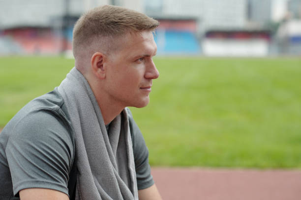 Profile of young athlete sitting on stadium track and gazing in distance. Man displaying contemplative expression, wrapped in grey towel with blurred stadium background