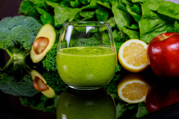 Fresh green smoothie with avocado, apple, and lemon on reflective surface surrounded by greens