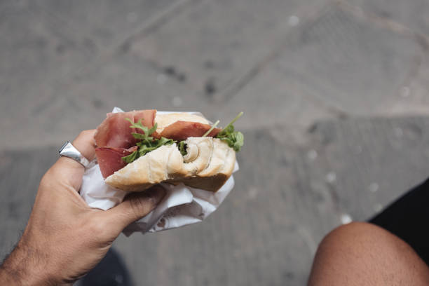 A close-up of a hand holding a fresh Italian panino filled with prosciutto and arugula, wrapped in paper, against a rustic street background in Florence, Italy. This simple yet flavorful sandwich highlights the essence of Italian street food culture.