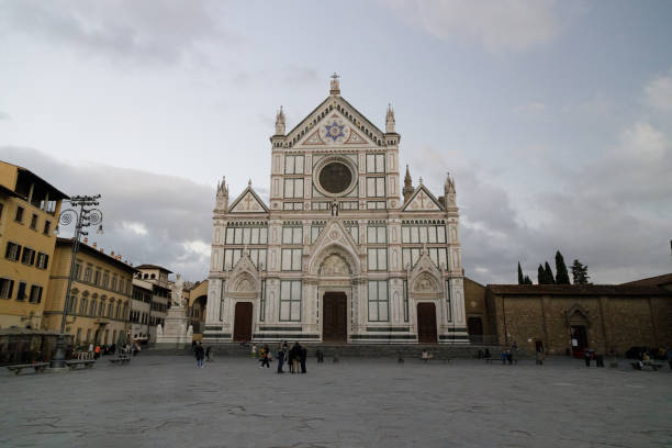 The Basilica of Santa Croce stands proudly in Piazza Santa Croce, showcasing its iconic Gothic architecture. The detailed façade, with its green and white marble, contrasts beautifully against the cloudy sky. The surrounding square, though sparse with people, evokes a serene and timeless atmosphere, inviting visitors to pause and reflect on the grandeur of this historic church. The sculpture in front adds a touch of artistry to the scene, complementing the rich heritage of this Florence landmark.