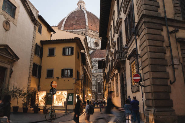 The evening light casts a soft glow on a narrow street in Florence, where the iconic dome of the Duomo towers over the historic buildings. The warm yellow light from the shops contrasts with the cool evening air, creating a charming atmosphere. People walk leisurely, some pausing to admire the beauty of the Duomo peeking through the alleyway. The sign marking a restricted street adds a sense of urban life, while the towering cathedral serves as a reminder of the timeless history and architectural grandeur of Florence.