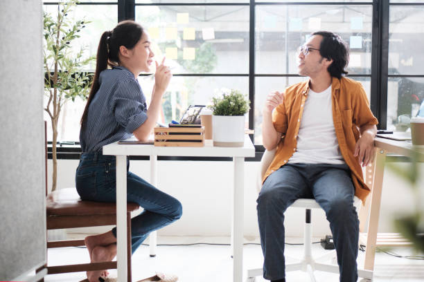 istock Two Asian workers at working desks near windows talk together. 2187073316