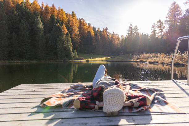 Woman in cozy attire lying on a blanket on a pier by a tranquil lake, soaking up the autumn sun with vibrant fall foliage and peaceful surroundings.