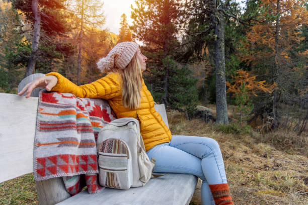 Woman in yellow jacket and beanie relaxes on a park bench in a forest. Casual and cozy atmosphere with autumn colors.