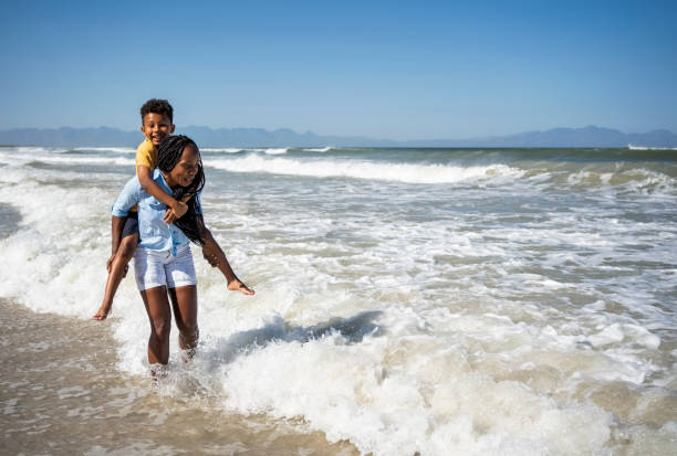 istock Young mother carrying son and walking on waves at the beach 2187075445