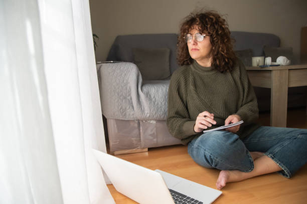 istock Focused Female Sitting On Floor With Laptop And Notepad, Working From Home In Natural Light. Mature Woman Near Window Writing Goals, Planning Her Day, And Organizing Agenda In Indoor Workspace 2187078852