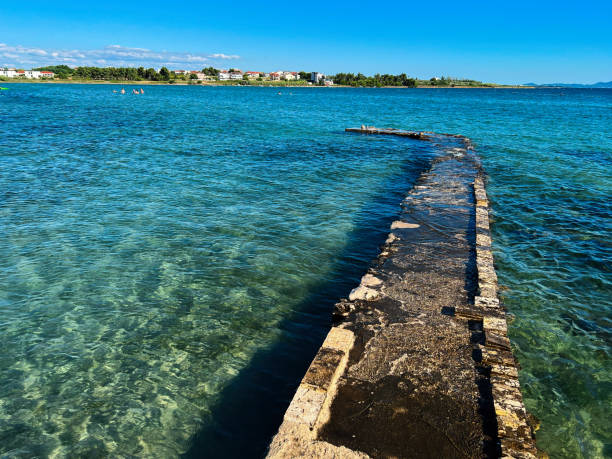 A serene coastal scene with a weathered stone path extending into the deep blue ocean. The calm water and clear sky create a peaceful, tranquil atmosphere perfect for relaxation.