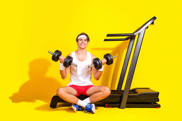 istock Young athlete in activewear smiling while lifting weights beside treadmill against bright yellow background 2187102635