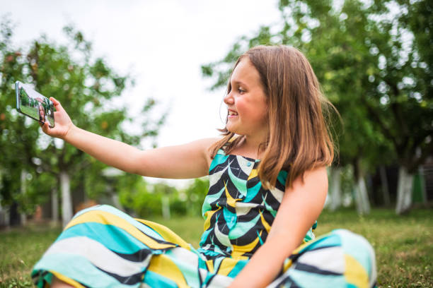 Girl sitting on green grass in a garden, taking a selfie on a sunny day, happy and content in nature with a smartphone. Reflecting modern youth's blend of outdoor fun and technology