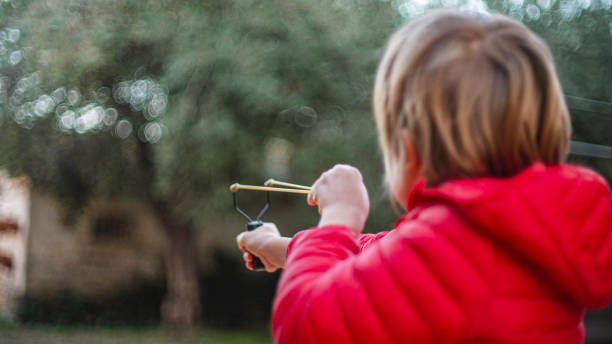 Child playing with a slingshot aiming at a flower pot in a garden stock photo