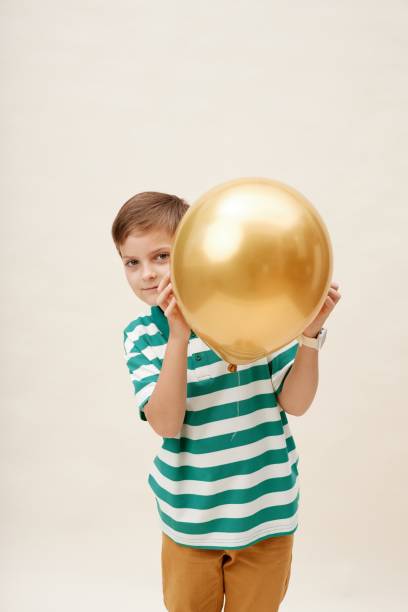 istock Smiling Boy Holding Yellow Balloon in Hands Hiding from Camera in Studio 2187109668