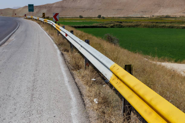 A highway stretches alongside lush green fields, featuring yellow guardrails and brown, arid mountains in the background