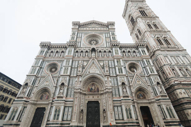 The magnificent Florence Cathedral, or Duomo, stands as a masterpiece of Gothic architecture, its intricate marble façade adorned with shades of green, white, and pink. Flanking it is the tall bell tower, designed by Giotto, which reaches toward the sky with its elegant proportions. At the center of this grand ensemble is the imposing entrance, complete with detailed statues and biblical scenes. The air is filled with reverence and history, as visitors marvel at the artistry and design that define this iconic cathedral in the heart of Florence.