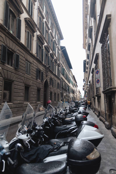 A quiet, rain-soaked street in Florence, with rows of scooters lined up neatly along the sidewalks. The buildings on both sides display the city’s characteristic blend of historical architecture and modern convenience. Despite the overcast sky, the scooters stand as a symbol of daily life in this bustling Italian city, where compact transportation is essential. The wet surfaces reflect the city’s rich textures, and the umbrellas held by pedestrians suggest the persistence of Florentine life even on a rainy day. This image captures a typical urban scene in the heart of Florence, where the old meets the new in a harmonious balance.