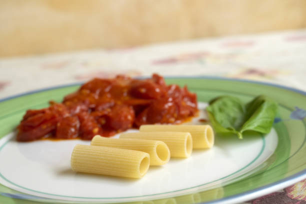 Stock photo showing close-up, elevated view of a red plate containing a meal of freshly cooked penne pasta tossed in a homemade marinara sauce made with tomatoes, garlic, olive oil, and herbs. The meal is topped with red cherry tomatoes, torn pieces of buffalo mozzarella cheese and basil leaves.