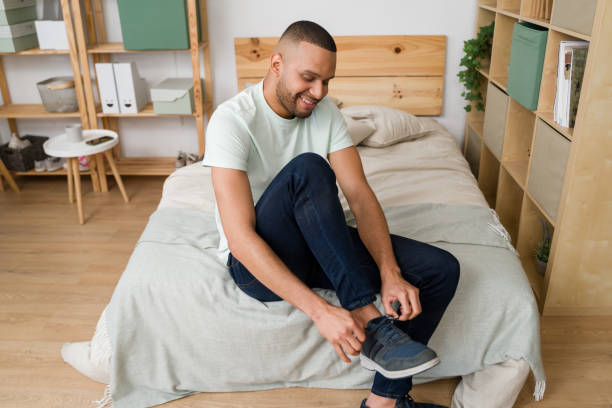 Young man tying his shoes while sitting on bed in his bedroom