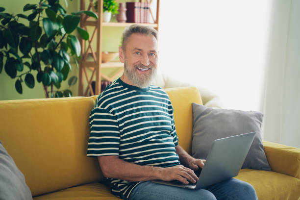 istock Elderly man enjoying a relaxing day at home using a laptop on a cozy yellow sofa in a bright living room 2187121378