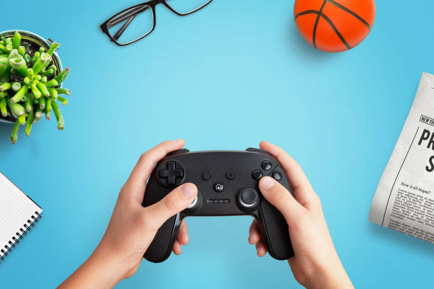 Top view flat lay composition of a player holding a gamepad on a desk with a ball, newspaper, glasses, plant, and notepad, perfect for creative gaming themes