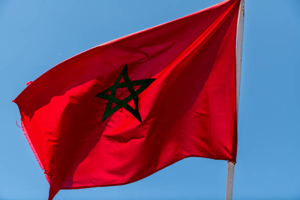 Moroccan flag fluttering in the wind with a red background and green pentagram, captured against a blue sky.