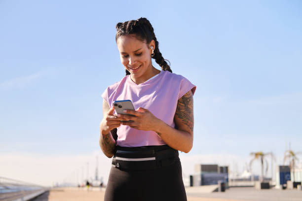 istock front view portrait of a fit and optimistic Latino female smiling using a smartphone  on a boardwalk in Barcelona city on a sunny day. technology and leisure activities concept 2187149350