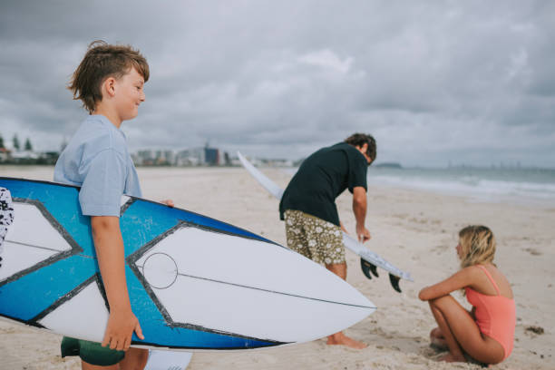 Portrait of a little boy stand with orange surfboard on the edge of the sea on the sand beach smile and hold the board