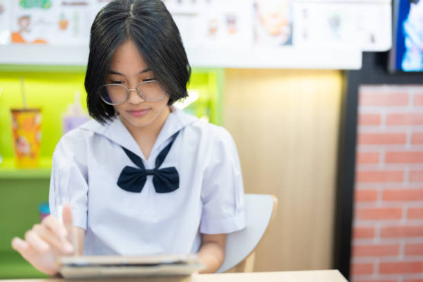 istock A diligent student in her school uniform is deeply engaged in writing on her tablet while sitting at a cozy and vibrant cafe, showcasing her focus and determination in learning. 2187174313