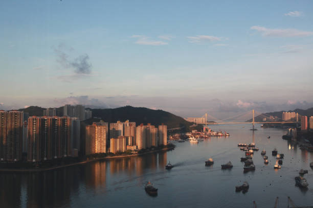 A bustling scene of Hong Kong at dawn. Towering residential skyscrapers reflecting the soft, golden hues of the morning sun. The high-rise buildings are tightly packed, emphasizing the city's dense urban landscape. Small boats and larger ships navigate the waterway, the background features faint outlines of distant hills, cloaked in a gentle morning mist that adds depth to the composition. The entire scene exudes a mix of serenity and energy, capturing the vibrant pulse of a city waking up to a new day.