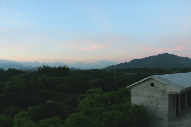 A picturesque village in China during the early morning. The sunrise bathes the sky in warm hues, with dramatic clouds hovering above distant mountains. Verdant greenery and simple village homes create a peaceful foreground.