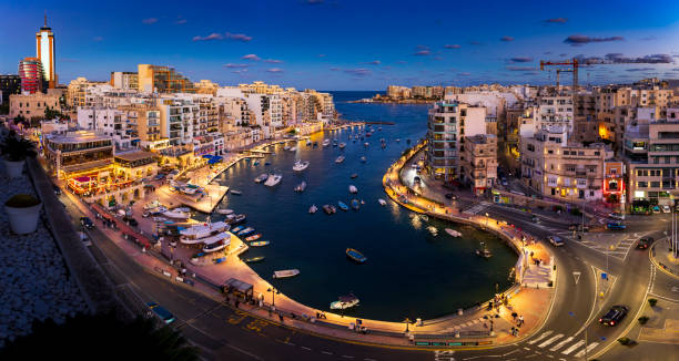 Elevated view of Spinola Bay in Malta at dusk, with illuminated buildings, boats on the water, and bustling streets.