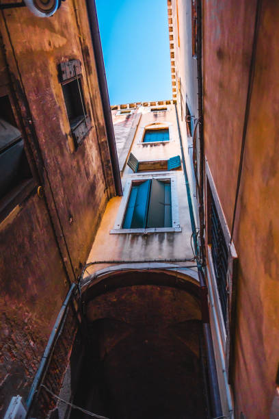 Looking up at a narrow Venetian alleyway, this image captures open shutters and rustic architecture against a vibrant blue sky, evoking a sense of history and charm.
