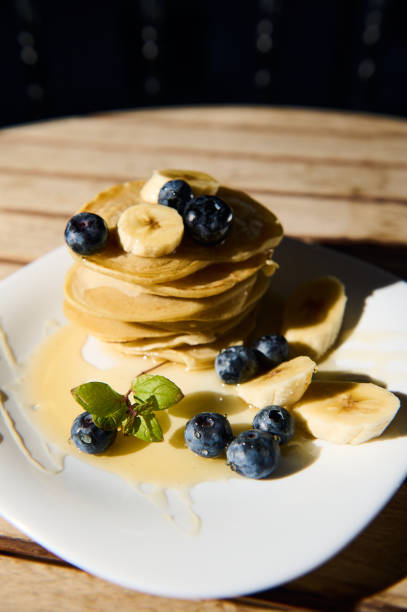 A tempting stack of golden pancakes topped with fresh banana slices and blueberries, drizzled with syrup on a white plate, capturing a delightful and sweet breakfast moment.