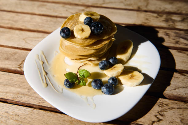 Delicious stack of pancakes topped with fresh banana slices and blueberries, drizzled with syrup. Perfect breakfast or brunch inspiration, showcasing fresh ingredients on a simple white plate.