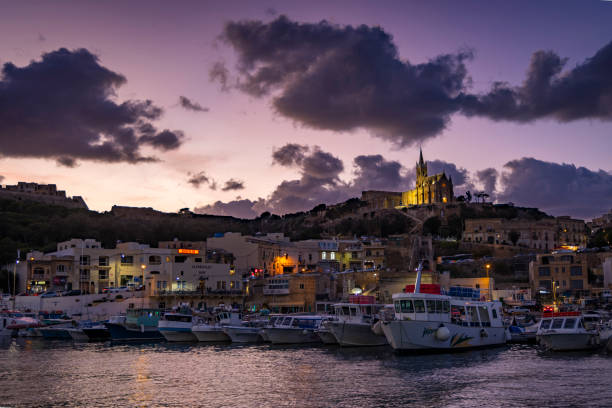 A vibrant sunset view over the port of Gozo, featuring illuminated buildings, docked boats, and the Church of Our Lady of Lourdes perched on a hill in the background.