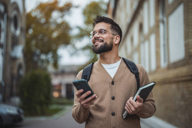 istock Smiling young student with backpack walking in front of university building and using mobile phone 2187202655