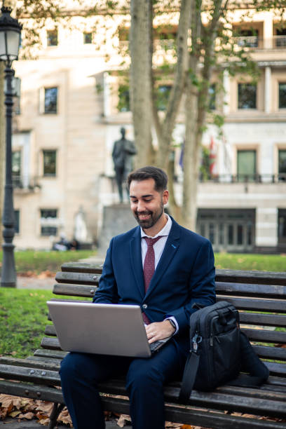 vertical businessman working on laptop outdoors at park bench with briefcase nearby