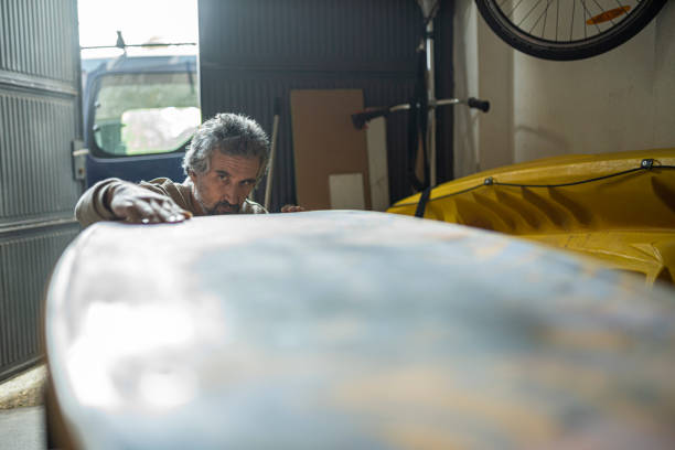 Green Business. Mature man repairing surfboard surface dings, searches for damage to the shell or core. Surfboard repair technician day in the life.