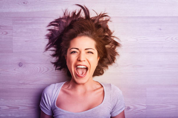 Young woman with messy brown hair lies on a wooden floor, her head tilted back and mouth wide open in a scream of frustration, joy, or excitement