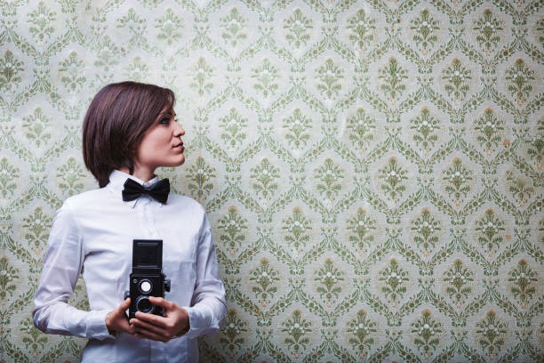 Young female photographer with bow tie and white shirt holding a vintage twin-lens reflex camera while looking sideways in front of patterned wallpaper