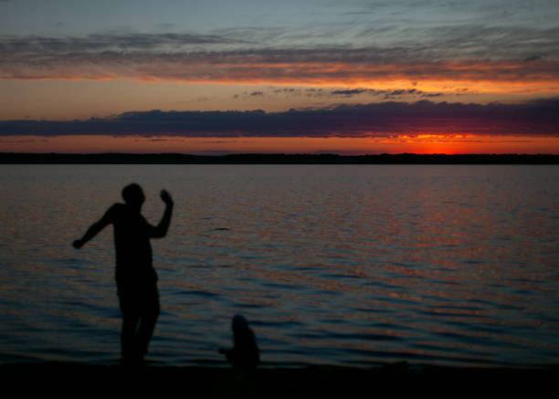 A peaceful silhouette of two figures, an adult and a child, enjoying time together by the lakeshore during a vibrant sunset. The fiery orange and purple tones of the sky reflect on the water, creating a tranquil and nostalgic atmosphere. Perfect for themes of family, connection, and serene outdoor moments.