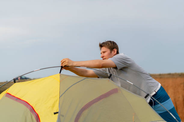 istock A man sets up a yellow tent on a grassy hill overlooking a lake. Setting camp during hiking. The sky is clear and blue, with some clouds in the distance 2187255046
