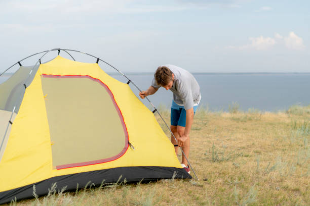 istock A man sets up a yellow tent on a grassy hill overlooking a lake. Setting camp during hiking. The sky is clear and blue, with some clouds in the distance 2187255054
