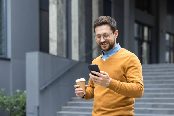 istock A smiling young male office worker went on a coffee break, standing outside the office building, holding a cup and using a mobile phone 2187294514