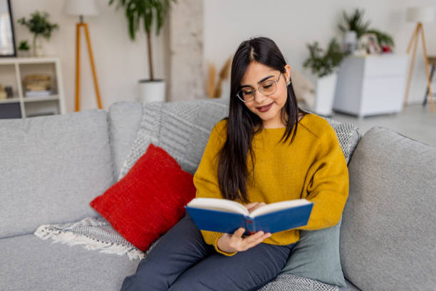 istock Dark-haired woman reading a book on gray sofa relaxes 2187295732