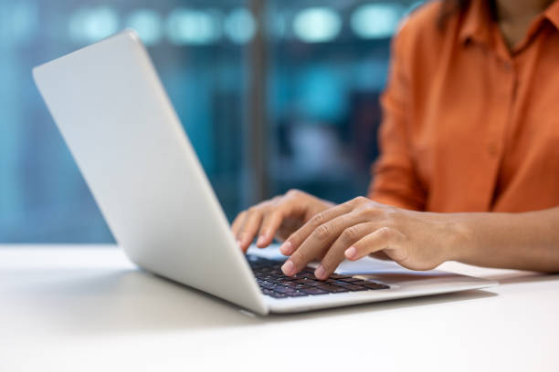 Close-up of hands typing on laptop keyboard in office setting, showcasing professional work environment. Focus on fingers pressing keys, dressed in business casual attire, reflecting modern work culture.
