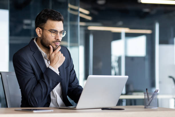 istock Focused businessman working on laptop in modern office setting, deep in thought, wearing glasses and a dark suit, reflecting a confident and professional demeanor 2187300342
