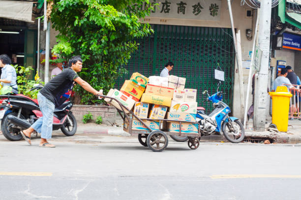 Thai man with pushcart  is transporting boxes over street Maha Rat Road in old Bangkok. Scene is near Wat Pho monastery. Man is passing old buildings with small stores of which one has closed jalousies. Man is wearing casual clothes. On sidewalk some people are walking,  One man is dressed like business  person with pants and necktie and has batch around neck. At right side people are stanbding at ATM