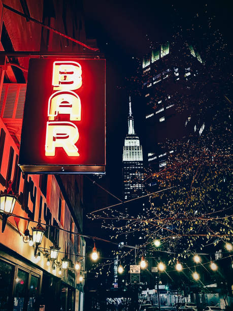 A vibrant red neon bar sign illuminates a nighttime city street, with the iconic Empire State Building visible in the background, creating a lively urban nightlife scene.