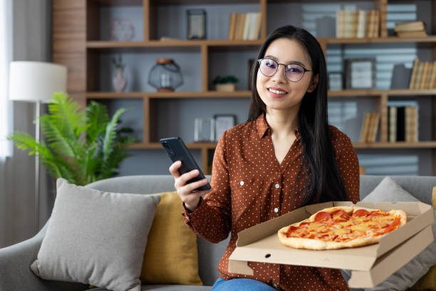 istock Smiling woman at home holding smartphone and enjoying pizza while sitting on cozy sofa, creating a relaxed and comfortable atmosphere. 2187306084