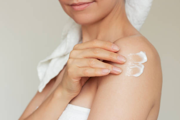 istock Woman wrapped in a white towel, smiling and touching her shoulder with her fingertips, applying a white cream 2187308932