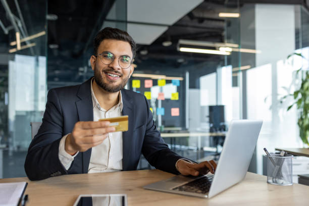 istock Smiling businessman using laptop holding credit card in modern office setting 2187310256